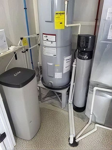 Residential utility room with water softener on left, central water heater with yellow maintenance tag, and water filtration system on right, connected by white pipes
