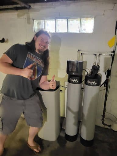 Person posing next to water filtration system tanks in an indoor utility room, giving thumbs up