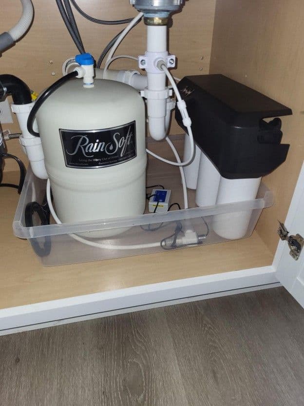 RainSoft water softener system installed under sink with white tank, filtration components, and connecting pipes in cabinet