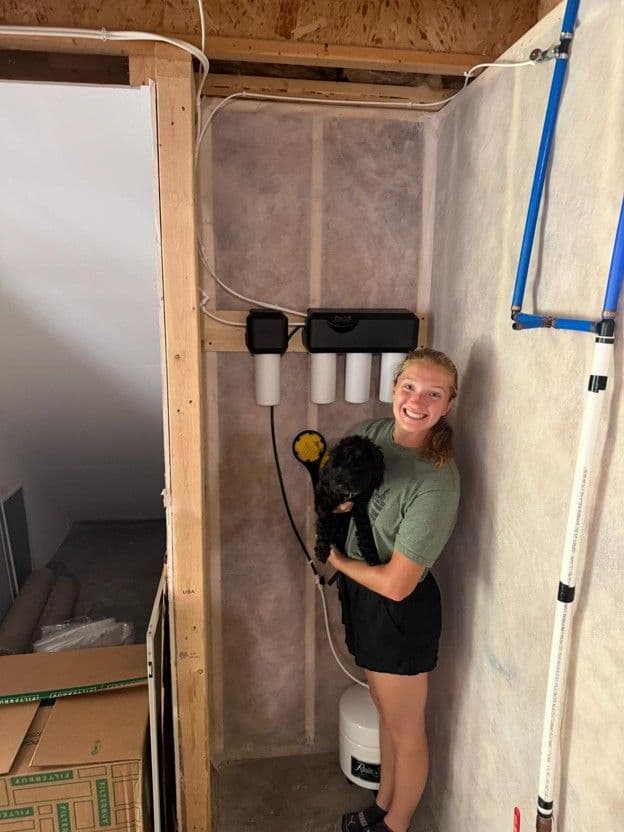 Woman holding black dog in unfinished bathroom under construction with exposed framing, drywall, and plumbing fixtures
