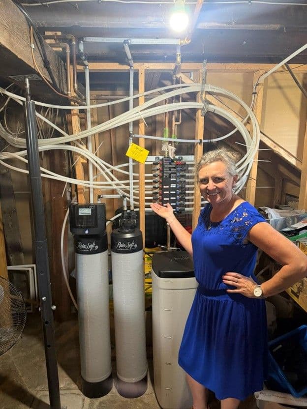 Woman in blue dress standing next to residential water softening and filtration system with white tanks and piping in basement utility room