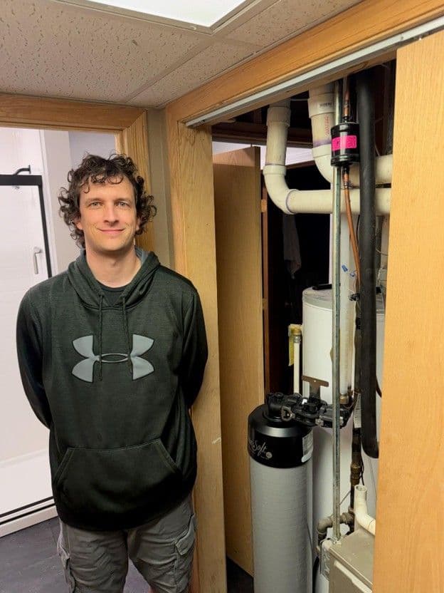 Man in Under Armour hoodie standing next to an open utility closet containing a water heater and plumbing pipes
