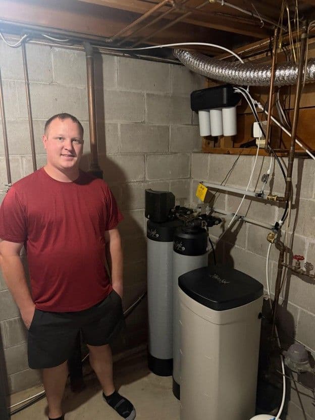 Man in red shirt standing next to water softening system with white tanks and piping in basement