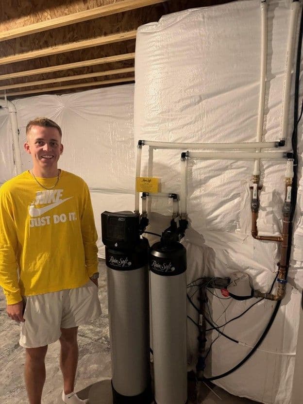 Man in yellow Nike shirt standing next to dual water softener tanks in basement with white plastic sheeting enclosure