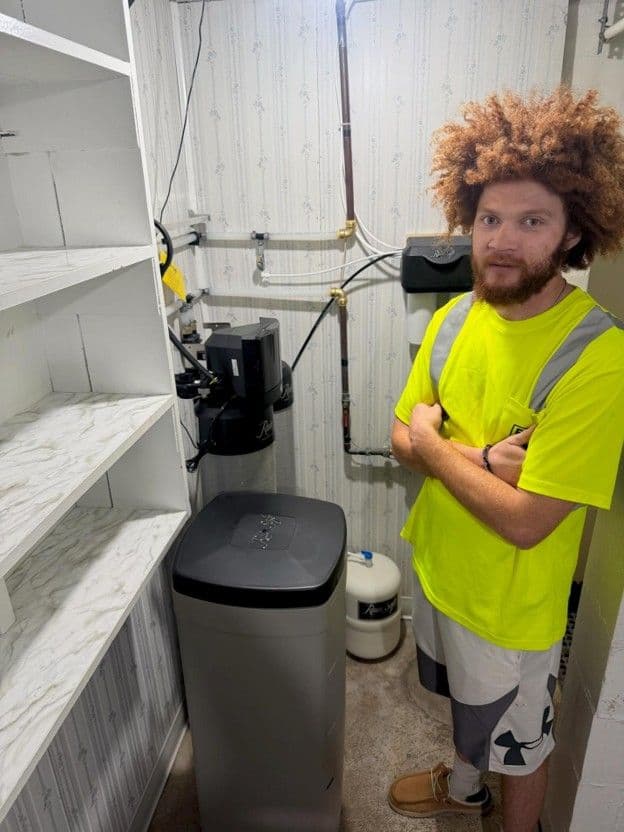 Plumber in high-visibility shirt standing next to installed water softener system with shelving and plumbing connections visible in utility room