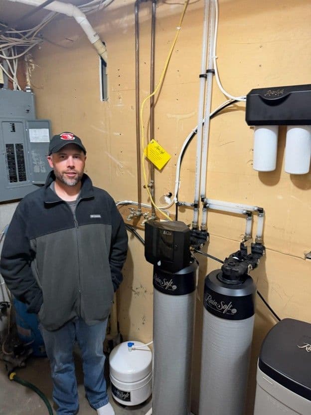 Technician standing next to installed water filtration system with dual tanks and connected tubing on yellow wall