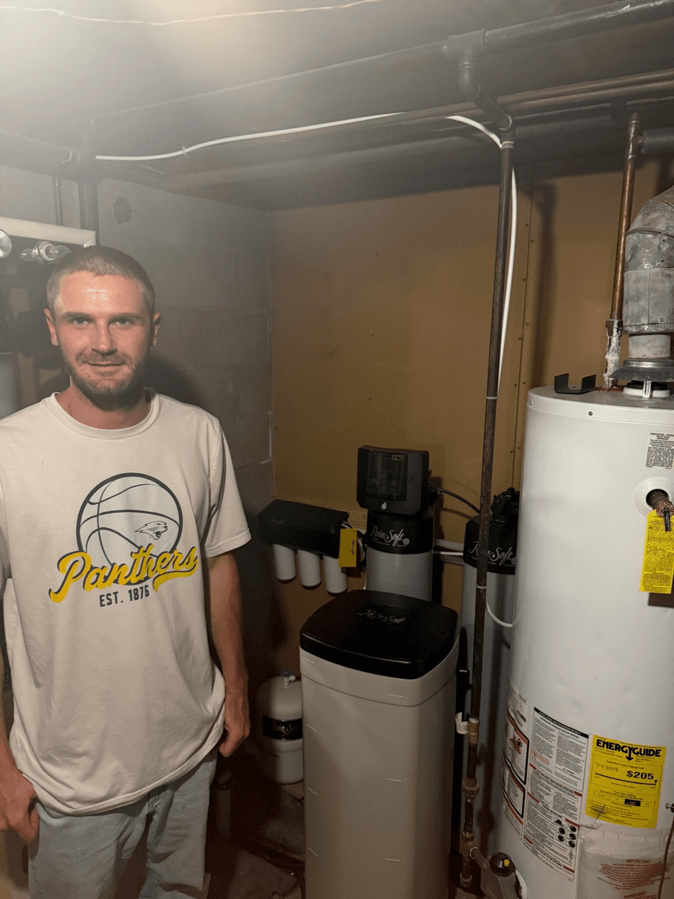 Man in Panthers t-shirt standing in basement utility room with water heater and water softener system