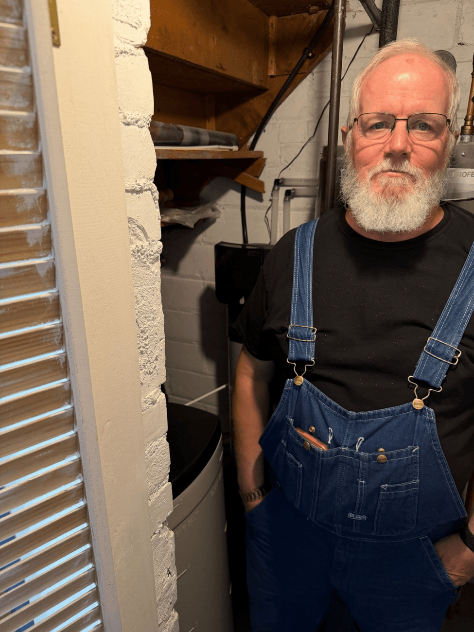 Man in denim overalls and glasses standing next to HVAC equipment and ductwork in basement utility room