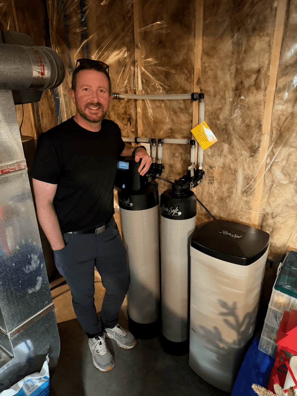 Man standing next to water softening system tanks in basement with exposed insulation on walls